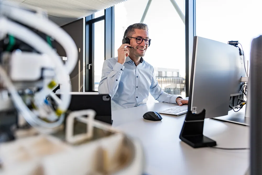 A man wearing glasses and a headset smiles while working at a computer in a modern office, with large windows and electronic devices on his desk.