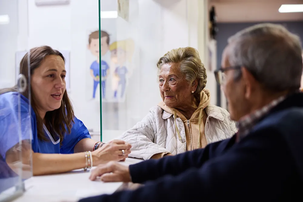 A nurse talks to an elderly woman and man across a clear partition at a medical reception desk. The elderly woman is wearing a light jacket and smiling, while the man sits beside her.