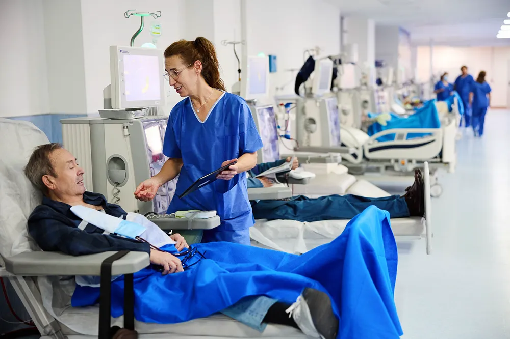 A nurse in blue scrubs stands beside a patient lying on a hospital bed connected to a dialysis machine, engaging in conversation in a medical treatment room.