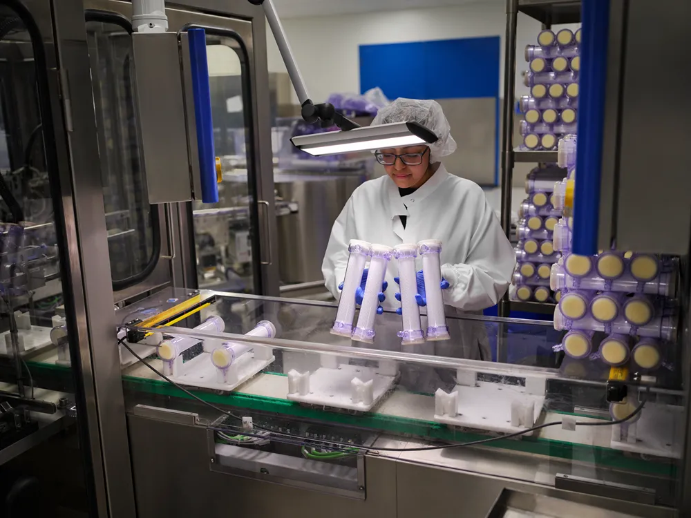 A person in protective clothing, gloves, and a hairnet inspects cylindrical medical devices under a bright lamp in a laboratory or manufacturing facility. Stacks of similar devices are visible in the background.