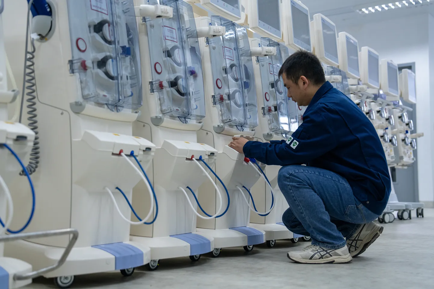 A man in a blue jacket and jeans kneels to inspect or adjust a row of medical dialysis machines in a clinical setting.