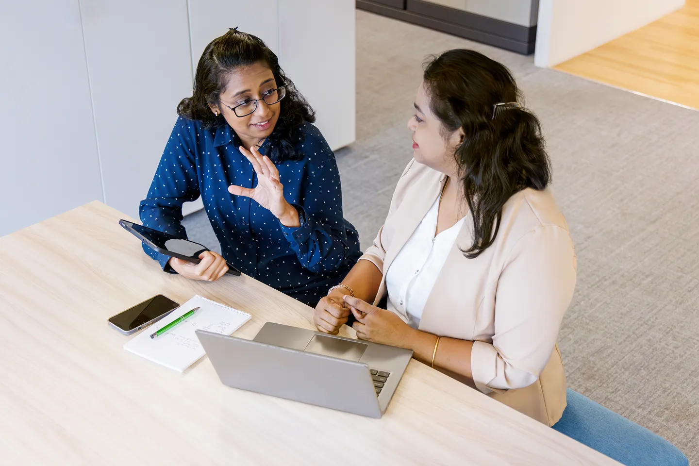 Two women sit at a desk having a discussion. One gestures while holding a tablet, the other listens. A laptop, notebook, phone, and pen are on the table. They appear to be in an office setting.