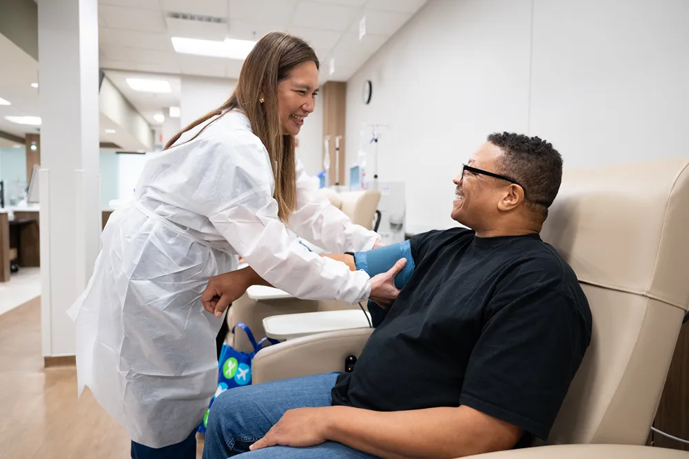 A healthcare worker in a white coat smiles while taking a seated man’s blood pressure in a medical facility. The man smiles back, and the setting appears clean and modern.
