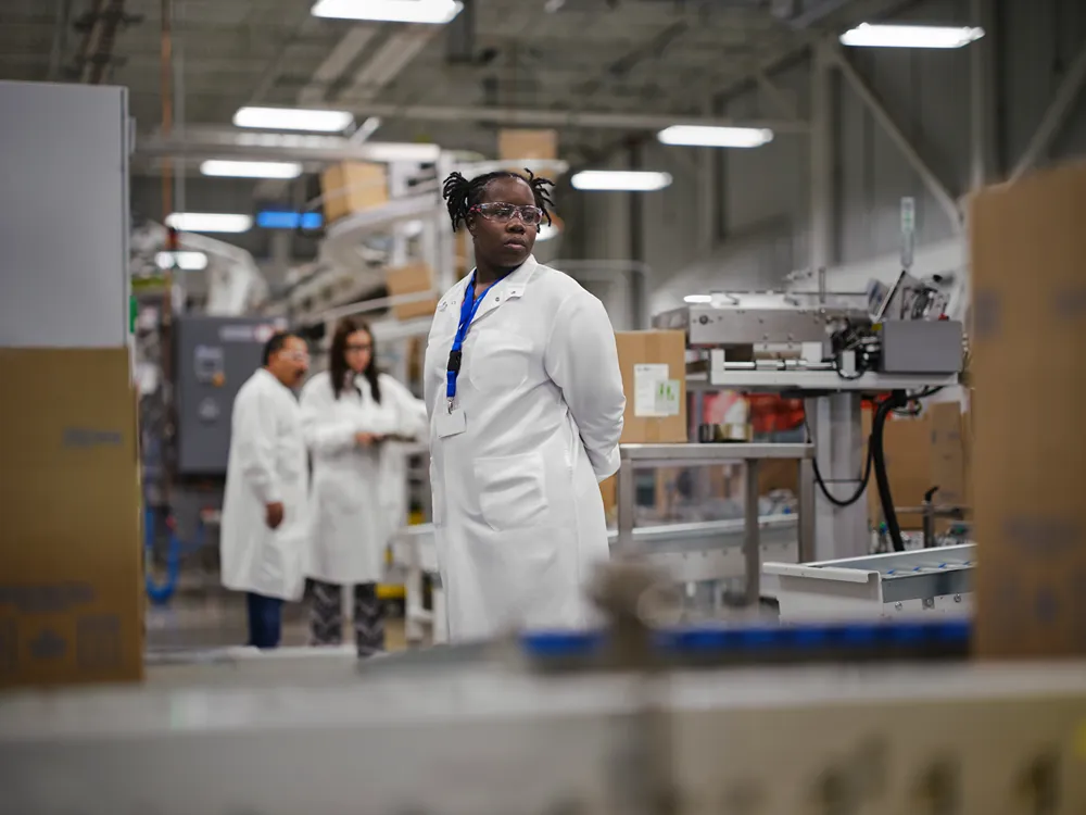 A person in a white lab coat, safety glasses, and blue lanyard stands in a factory or laboratory setting, with machinery and three other people in lab coats visible in the background.