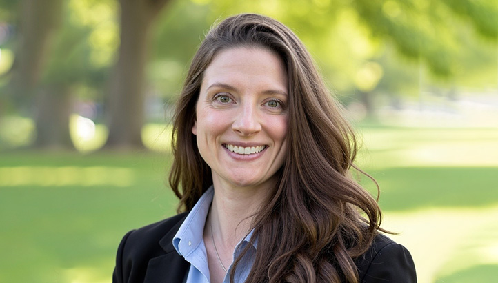A woman with long brown hair, wearing a black blazer and light blue shirt, smiles outdoors with green grass and trees in the background.