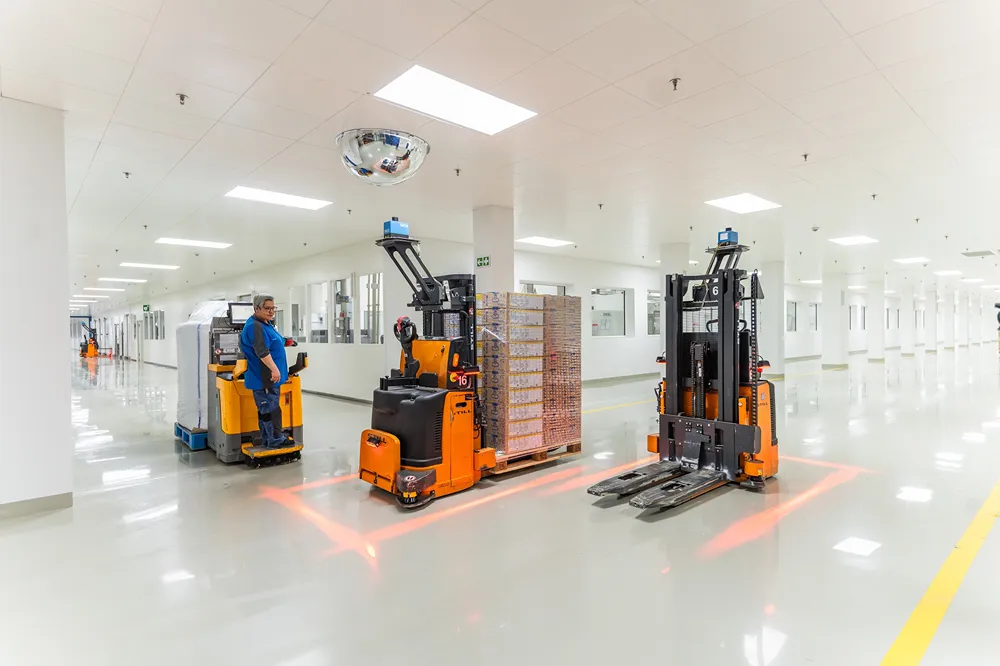 A worker in blue operates a vehicle beside two orange autonomous forklifts carrying metal racks in a clean, bright, industrial facility with white walls and shiny floors.