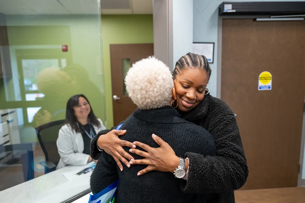 Two women warmly hug each other in a clinic, one facing the camera with a smile, while a woman in a white coat sits behind a glass window, also smiling.