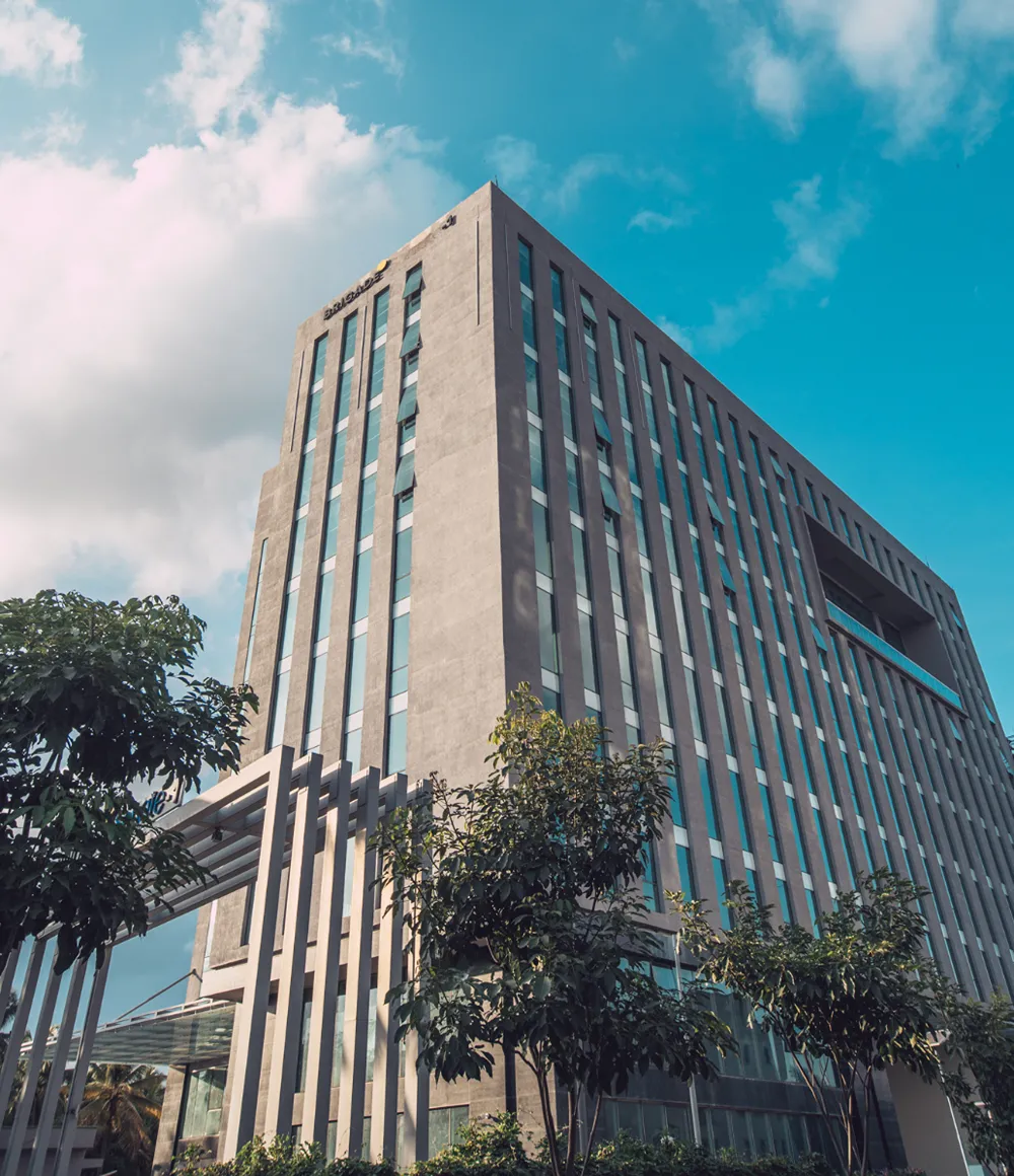 A modern high-rise building with vertical glass windows and concrete panels, surrounded by green trees, set against a blue sky with some clouds.