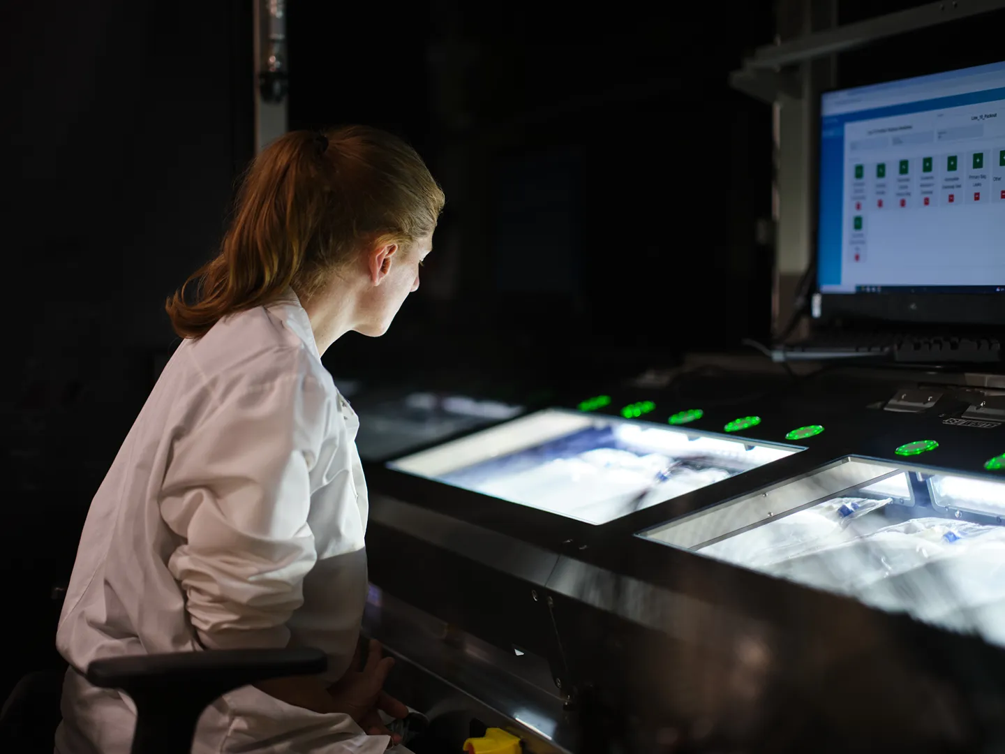 A person in a white lab coat sits at a workstation with illuminated surfaces, looking at screens with data or images displayed in a dimly lit laboratory or research environment.