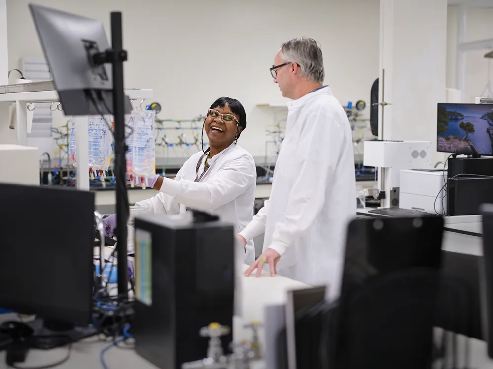 Two scientists in white lab coats smile and talk while working in a laboratory with computers and scientific equipment in the background.