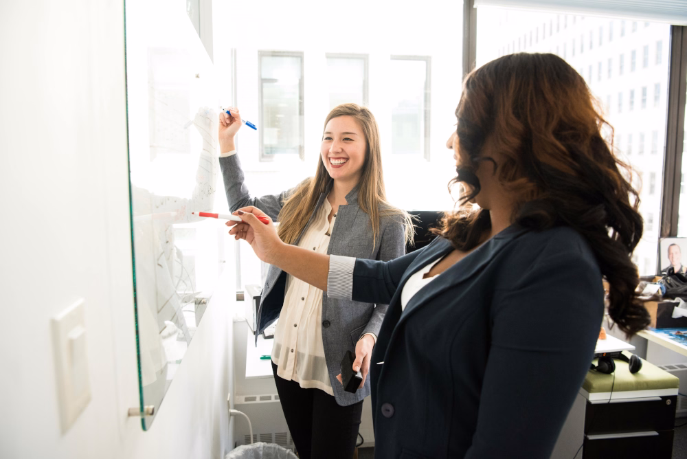 Two women in office attire writing and collaborating on whiteboard, smiling together.