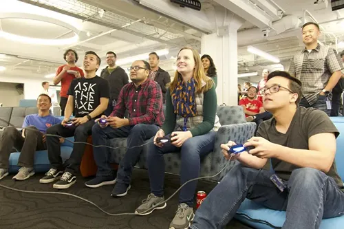 A group of people, including men and women, are sitting on couches in a room with white walls and exposed ceiling beams, playing video games using controllers. Some are laughing and smiling while others watch them play.