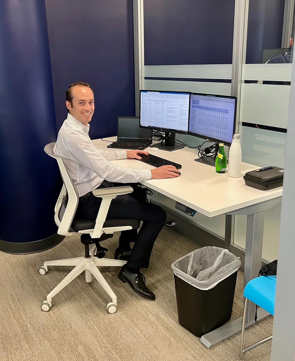 A smiling male professional sitting at an office desk with two monitors, looking at the camera