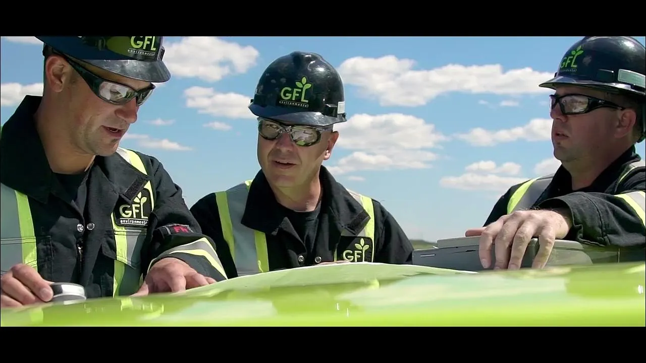 Three workers in GFL Environmental hard hats and safety glasses review documents outdoors