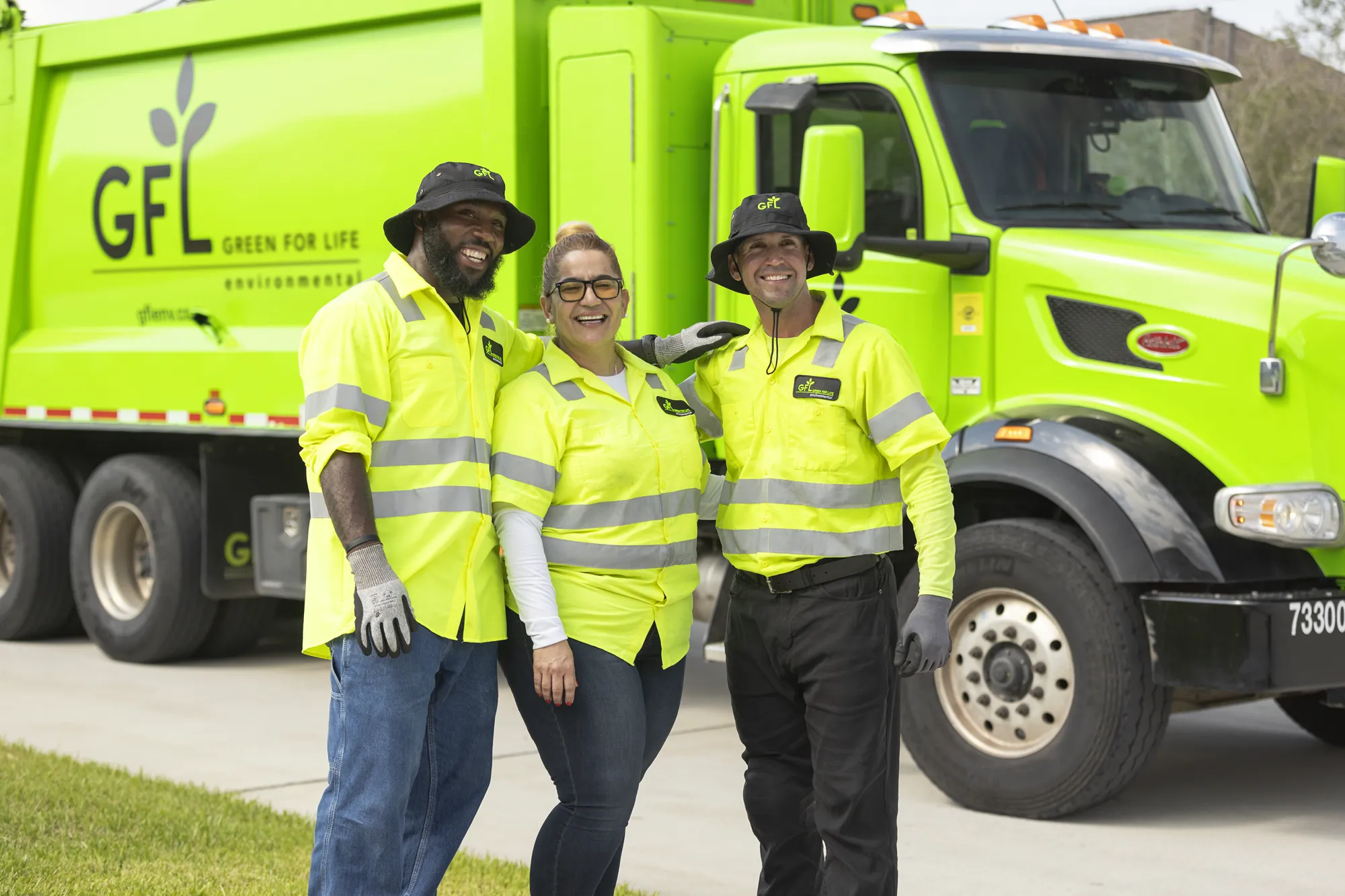 Three smiling Green For Life Environmental employees in bright yellow safety vests stand in front of a green garbage truck