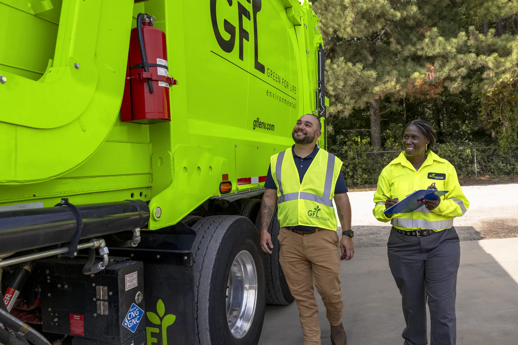 Two Green For Life Environmental employees walk beside a bright green garbage truck