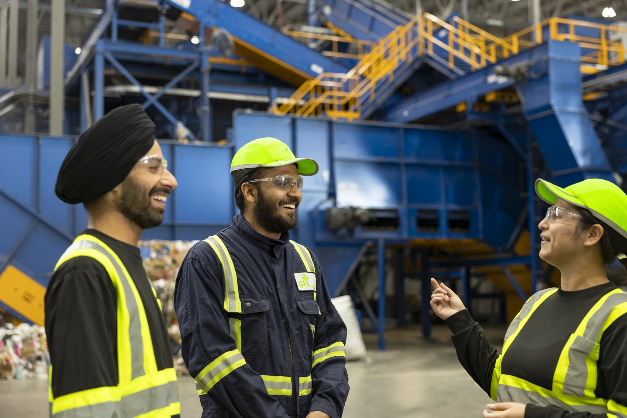 Trois ouvriers souriants en équipement de sécurité conversent dans une usine de recyclage.