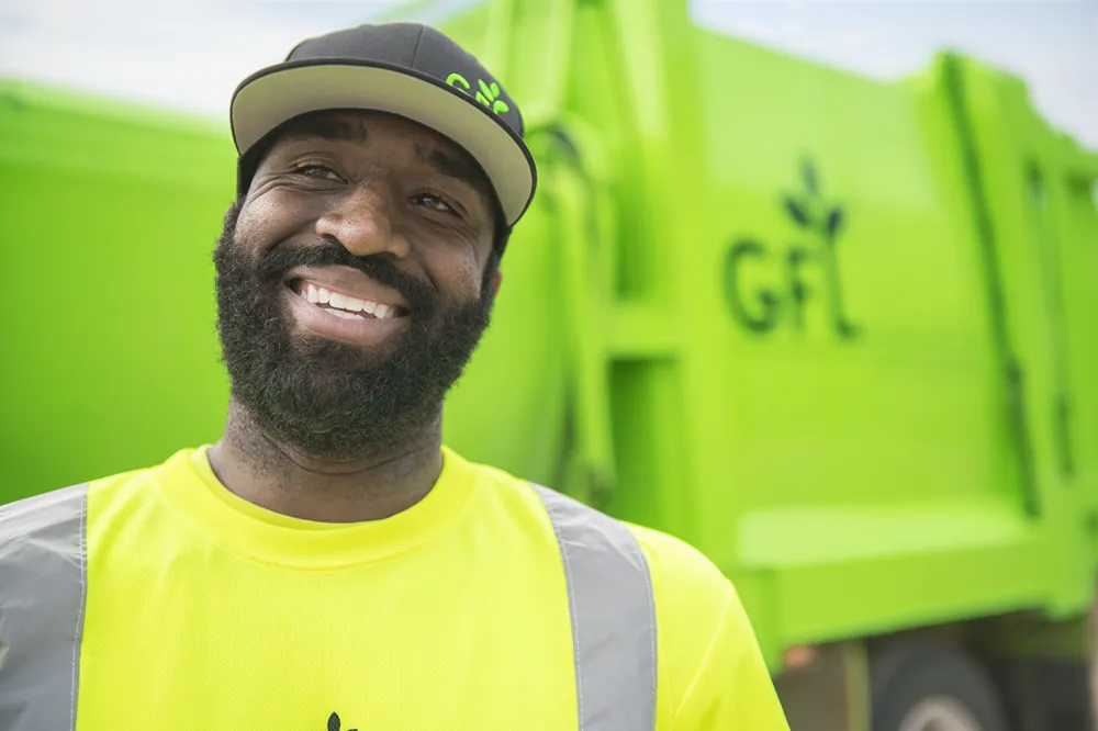 A smiling man in a yellow shirt and safety vest stands in front of a green garbage truck, representing community service and giving back