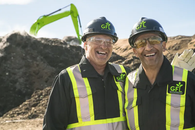 Two smiling men in GFL Environmental hard hats and safety vests stand in front of a pile of dirt and an excavator