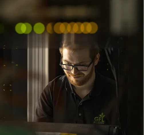 Un homme avec des lunettes et une barbe regarde un ordinateur dans une salle de serveurs faiblement éclairée.