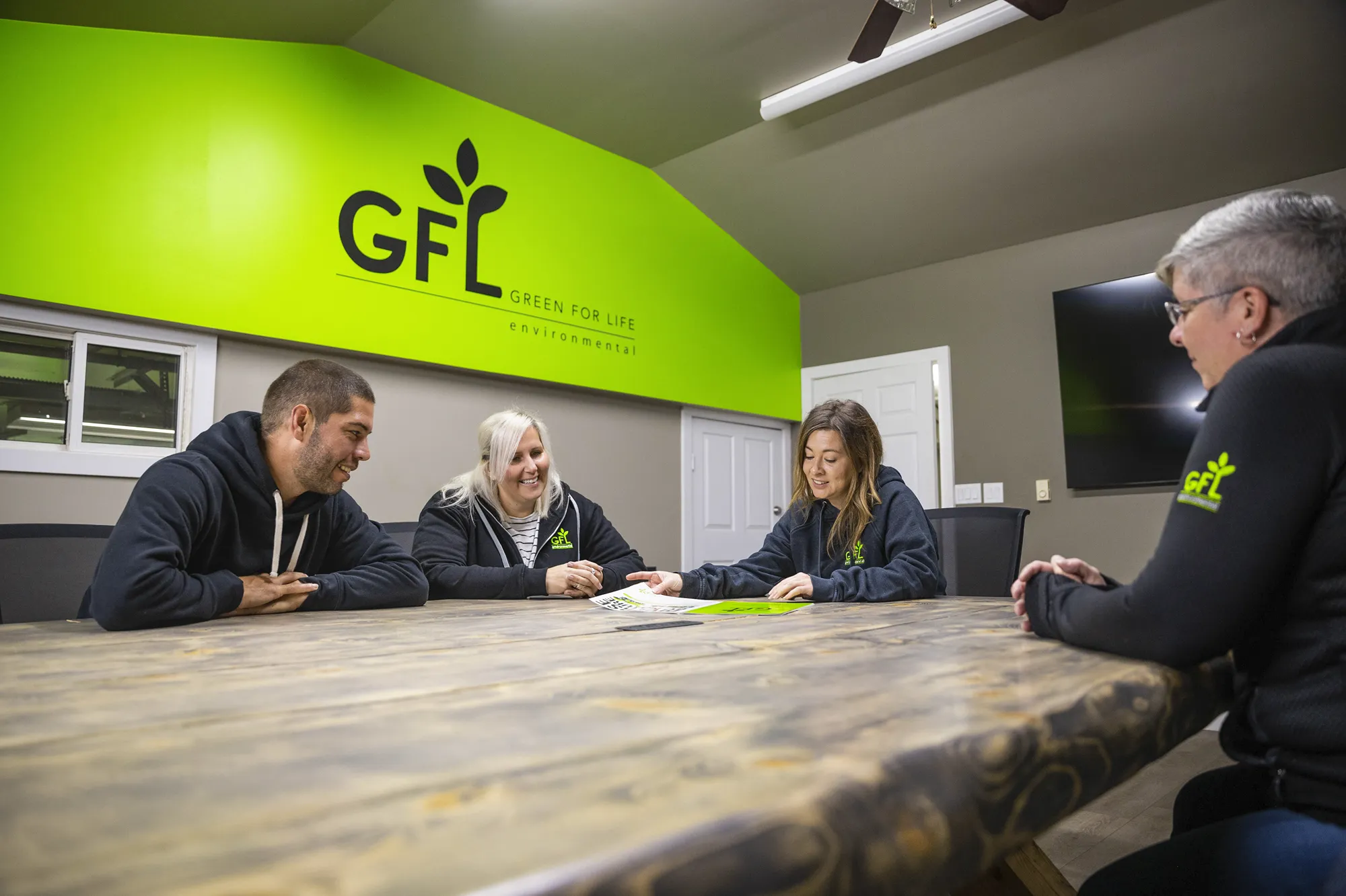 Quatre personnes sont assises autour d'une grande table en bois, examinant des documents, dans une salle de réunion avec un mur vert vif arborant le logo Green For Life Environmental.