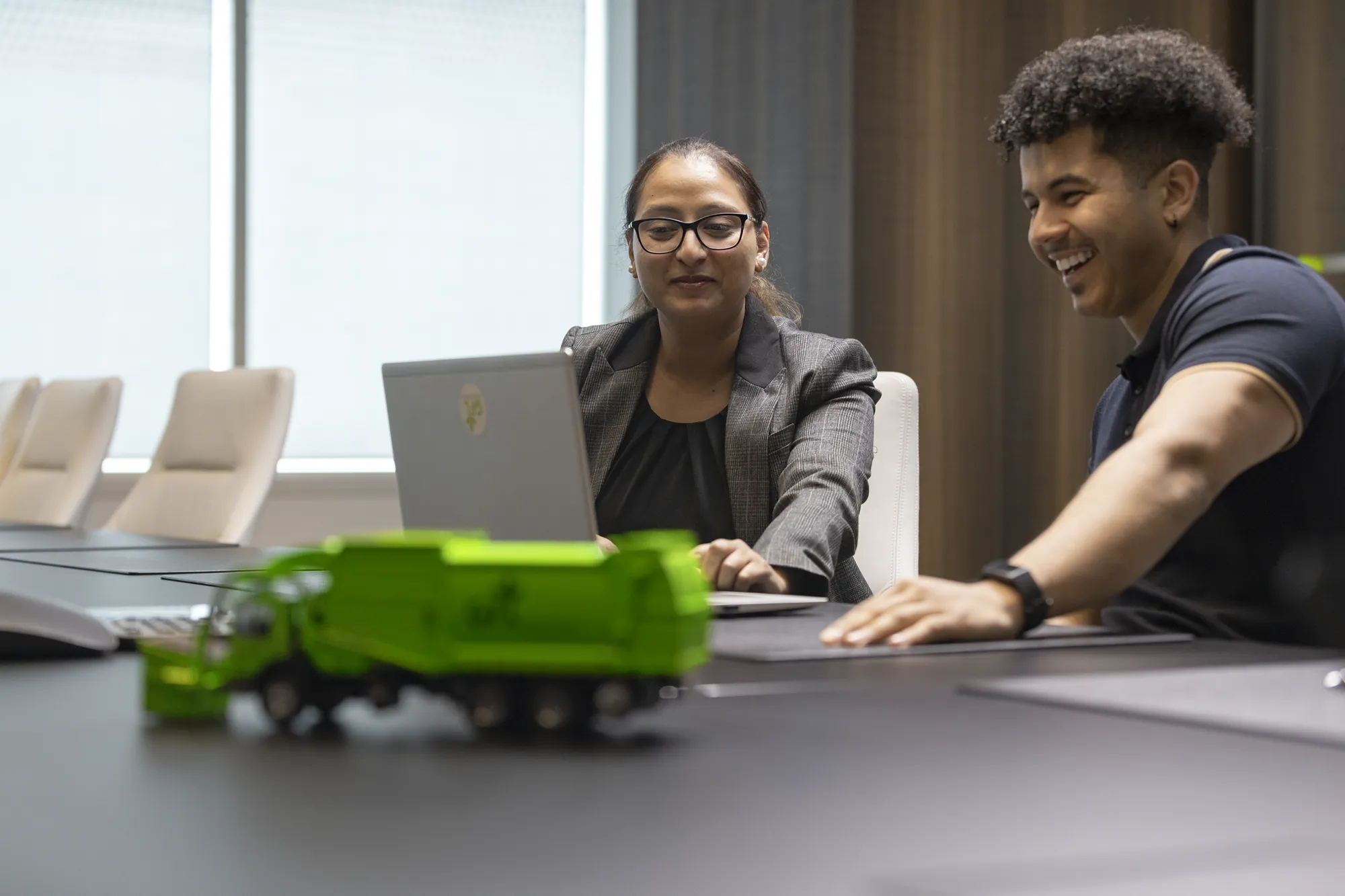Deux collègues collaborent sur un ordinateur portable dans un bureau moderne, un camion à ordures jouet au premier plan.