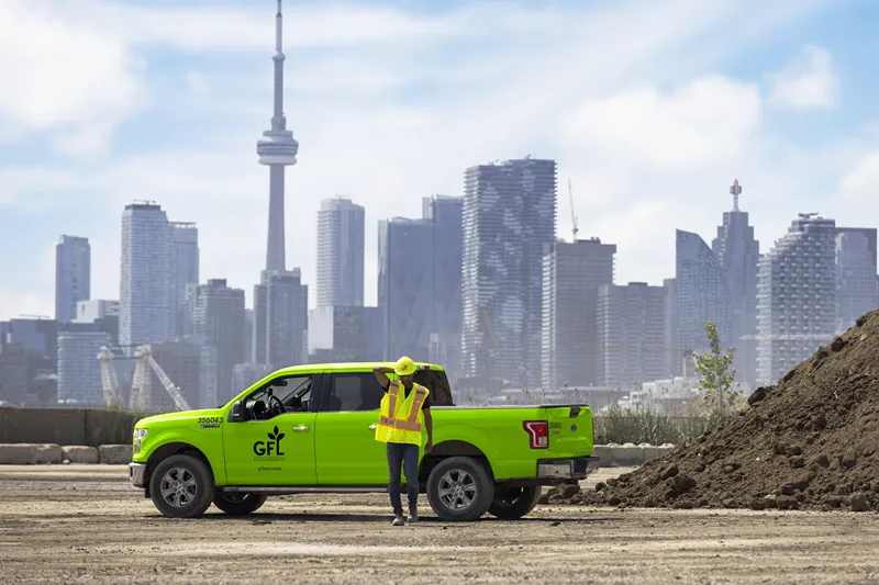 A worker in a safety vest stands by a bright green GFL Environmental truck in front of the Toronto skyline