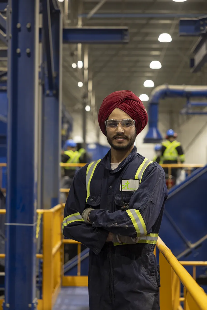 A man in a dark blue uniform and red turban stands with his arms crossed in an industrial setting