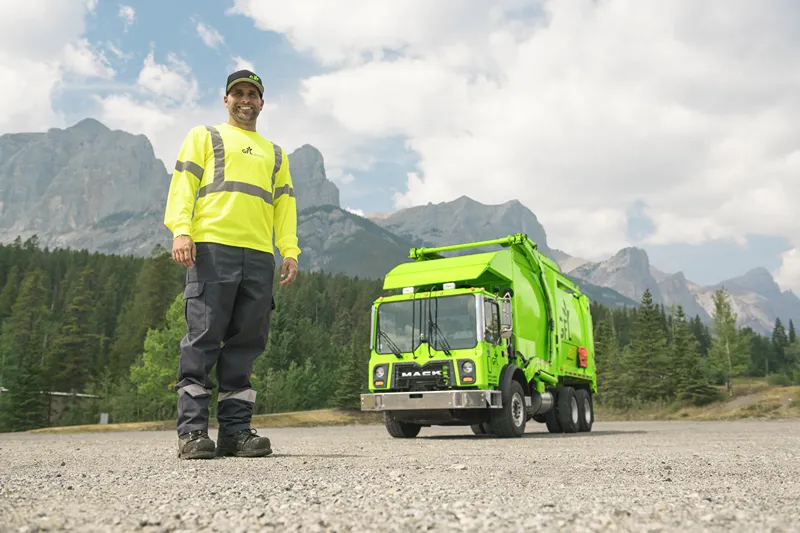 A smiling sanitation worker in a bright yellow uniform stands in front of a lime green garbage truck in a mountainous landscape