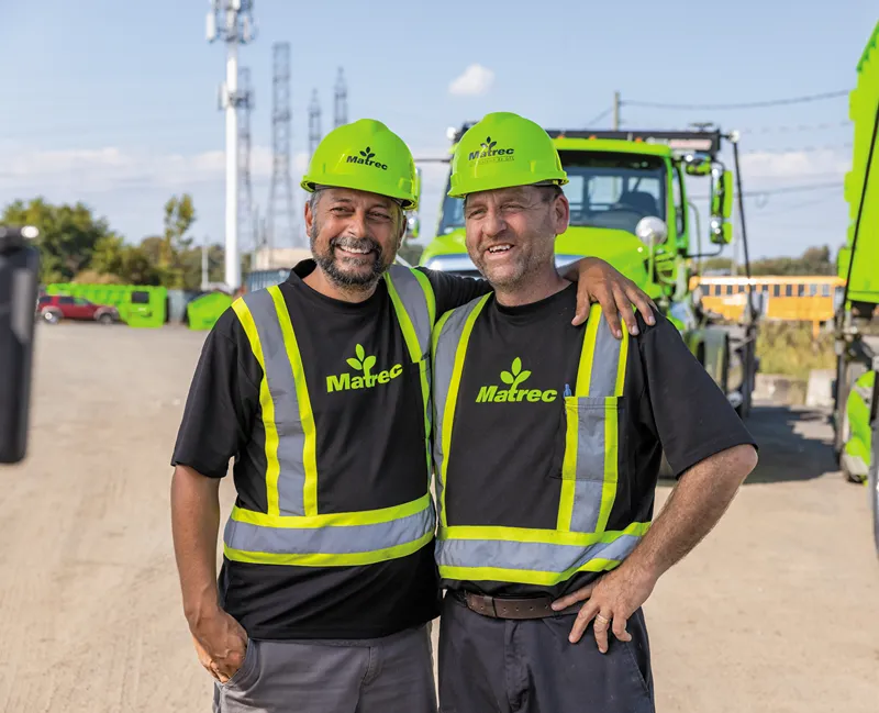 Deux hommes souriants portant des casques et des gilets de sécurité GFL Environnement se tiennent devant un tas de terre et une pelleteuse.