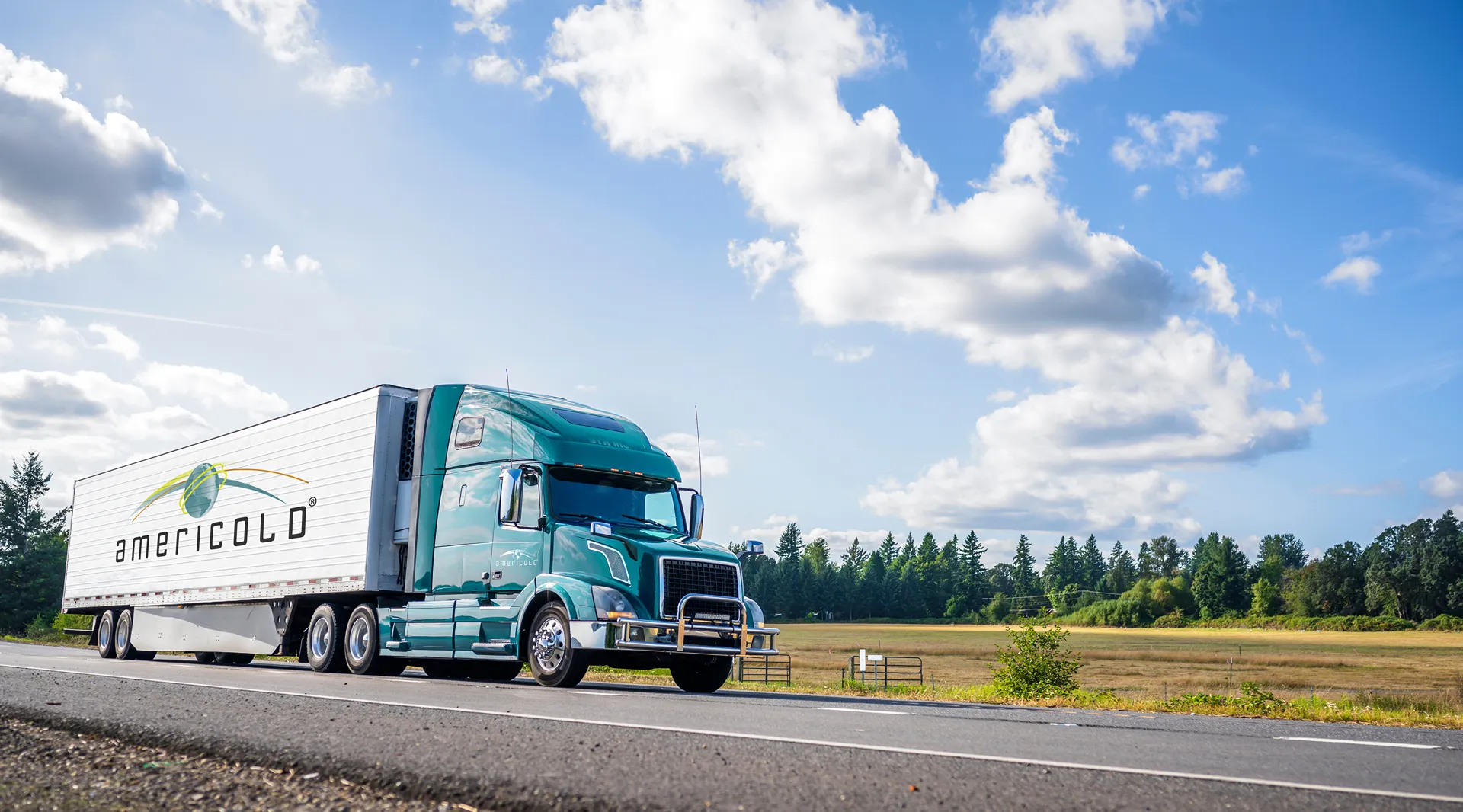 A green Americold semi-truck drives on a sunny road with fields in the background.