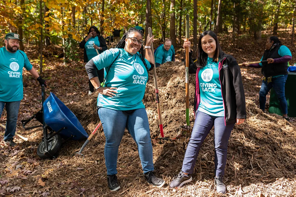 Volunteers in teal shirts work together to build a compost pile in a forest setting.