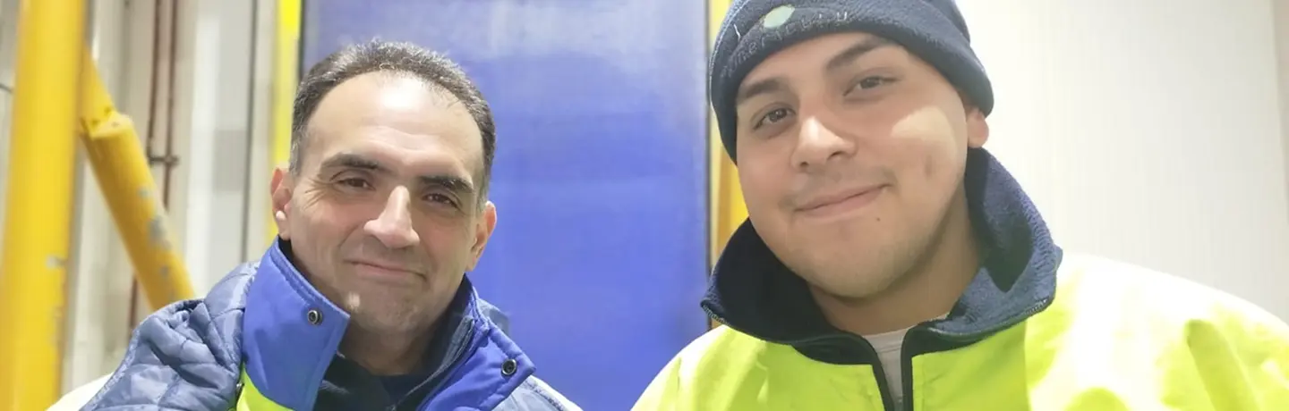 Two men in safety jackets smile at camera inside industrial setting
