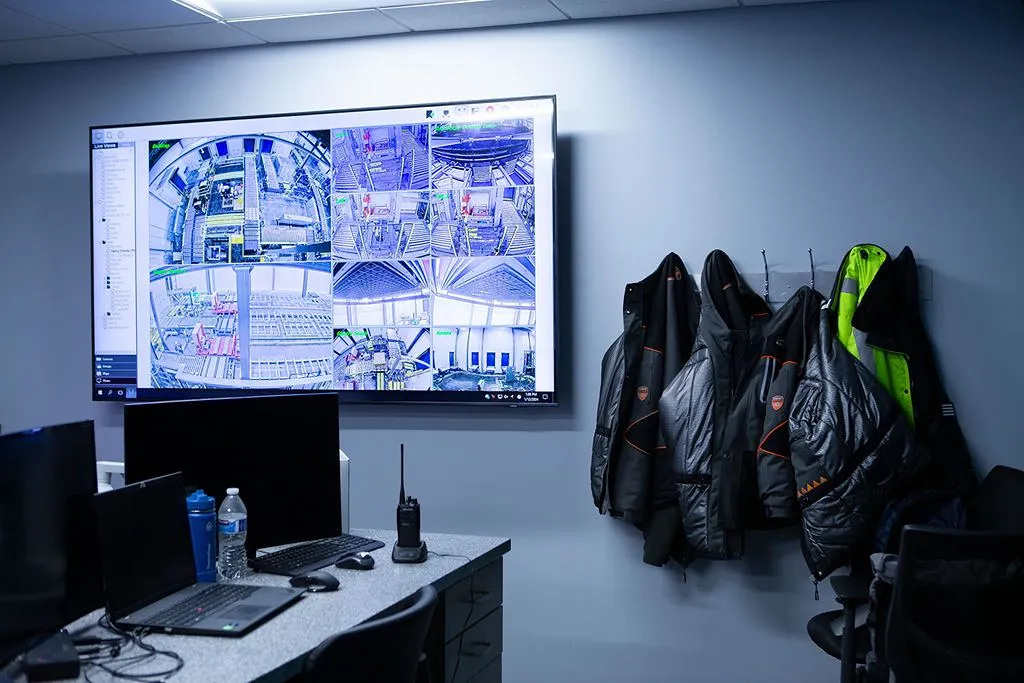 Surveillance monitoring room in a warehouse with large screen displaying multiple security camera feeds, workstations with laptops and radios, and safety jackets hanging on wall hooks.