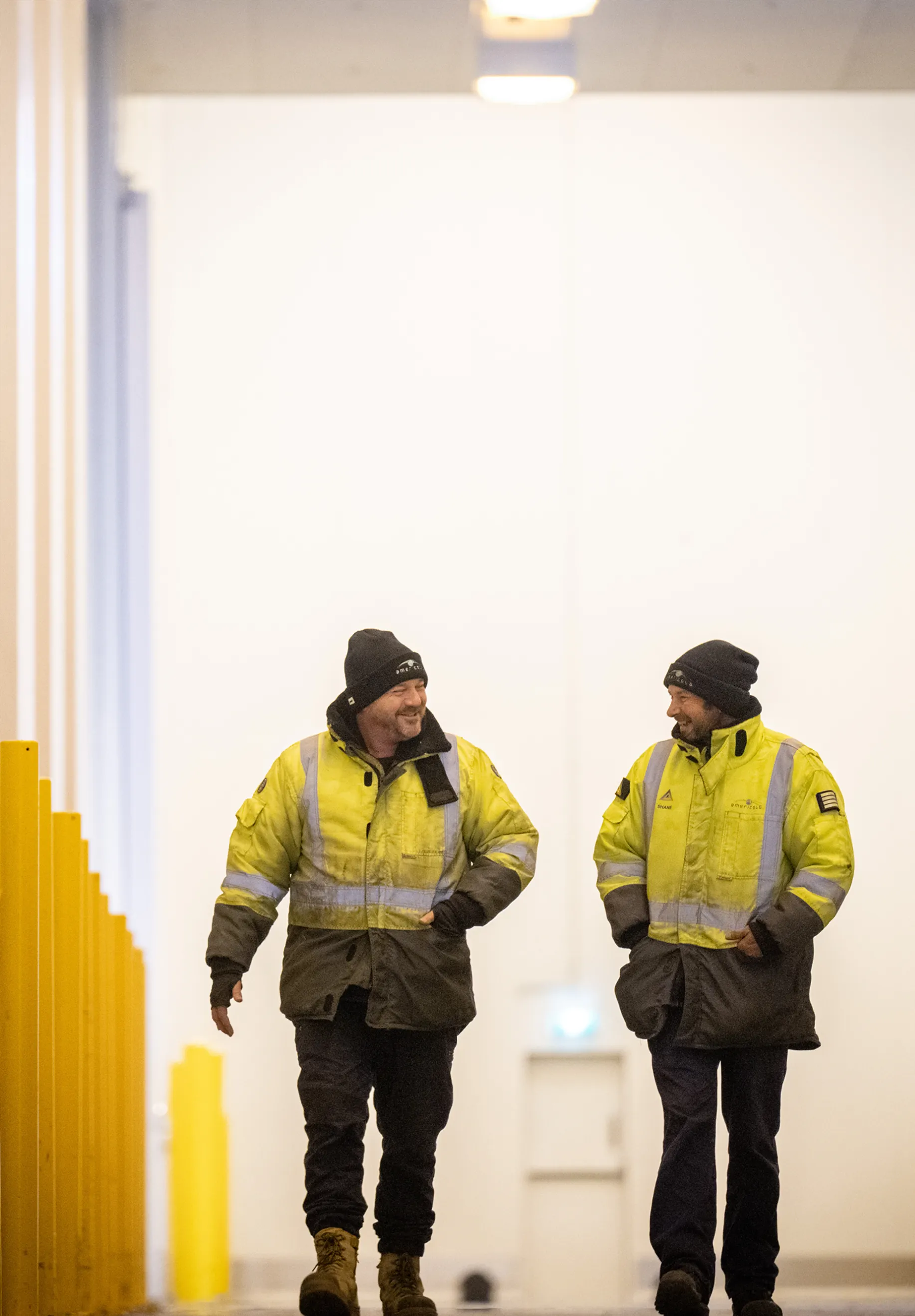 Two individuals in yellow safety gear walk through an industrial setting, engaged in conversation while navigating the space.