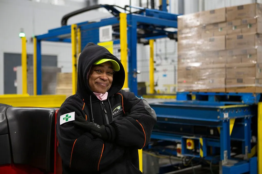 Trabajador de almacén sonriente con equipo de seguridad de pie con confianza cerca de maquinaria de embalaje automatizada con paletas apiladas en el fondo dentro de un centro de distribución