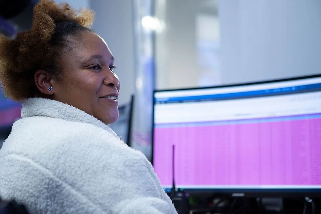 A person with curly hair wearing a white fluffy jacket sits in front of a computer monitor displaying a pink and blue interface, likely in a modern office setting.