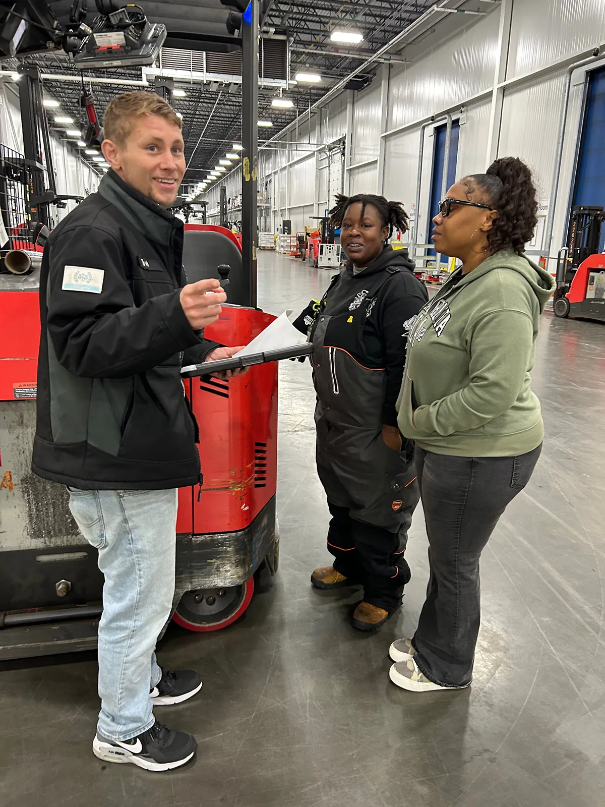 Three individuals are standing in a large warehouse with forklifts and industrial equipment in the background.