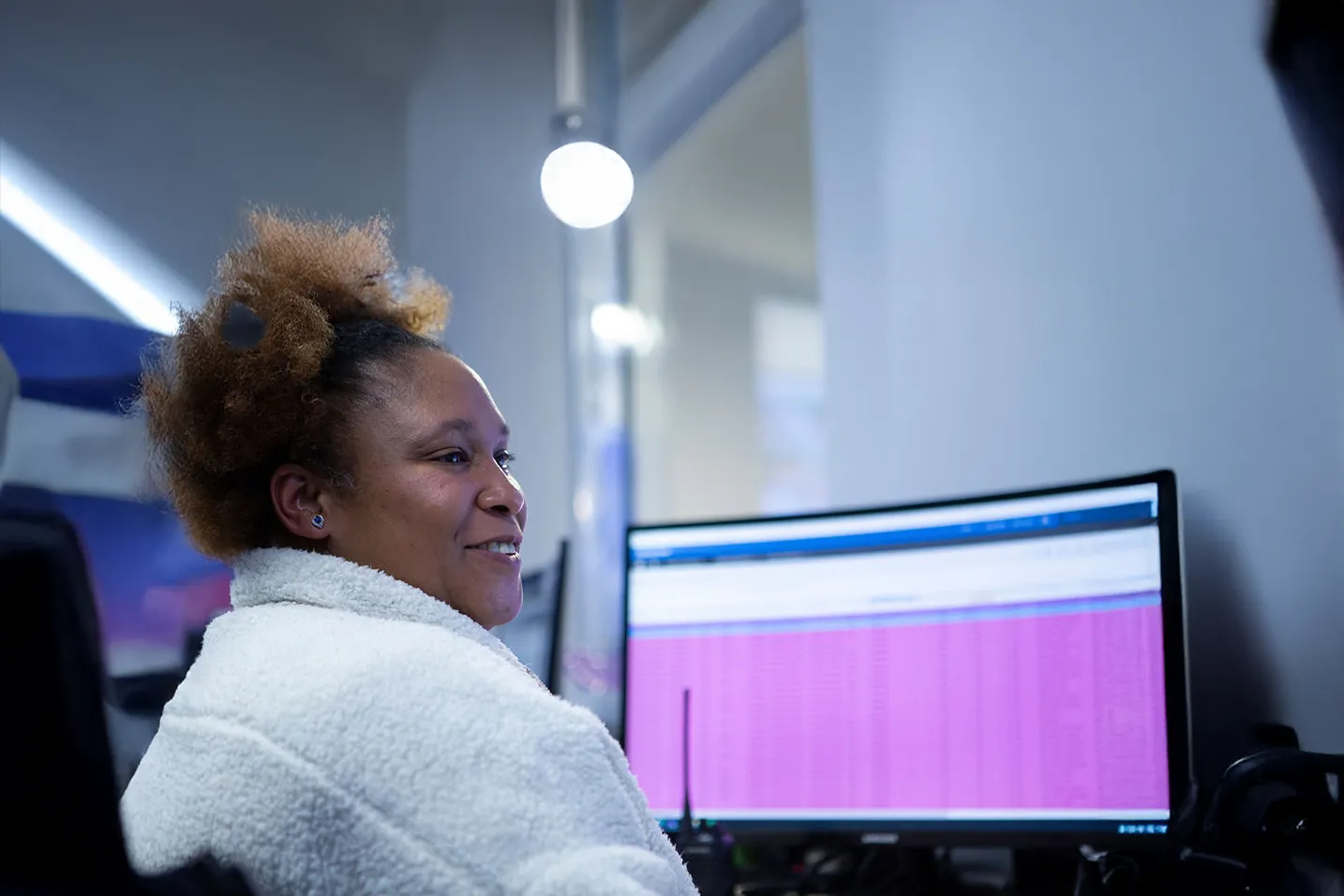 A person with curly hair wearing a white fluffy jacket sits in front of a computer monitor displaying a pink and blue interface, likely in a modern office setting.