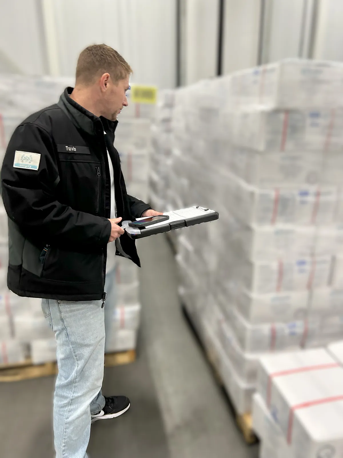 A person wearing a black jacket and light blue shirt stands in a warehouse, holding a clipboard and writing with a pen. The background features industrial equipment and a clean, organized interior.