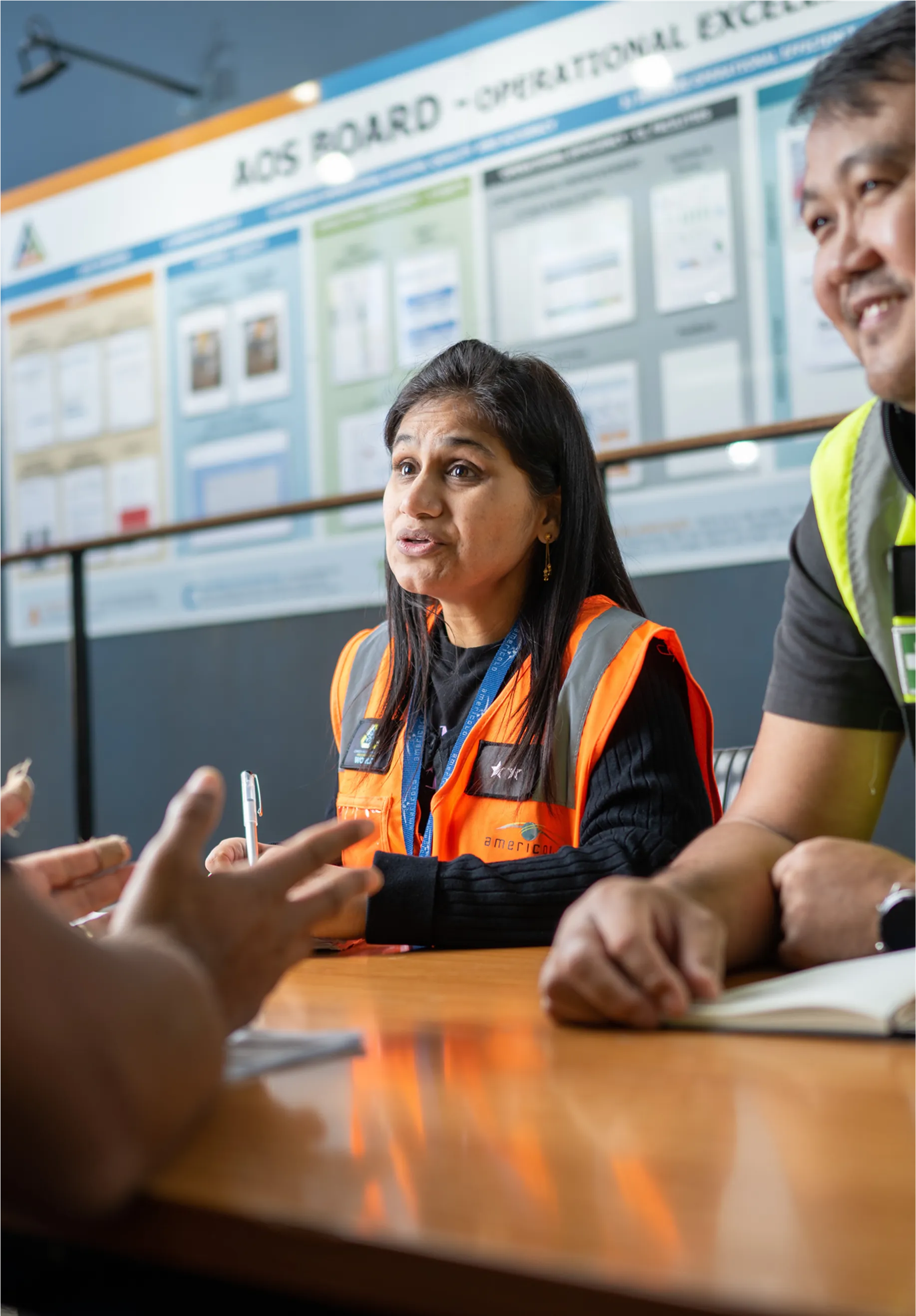 Three individuals in safety vests discuss at a table, with an `AOS Board - Operational Excellence` board behind them.