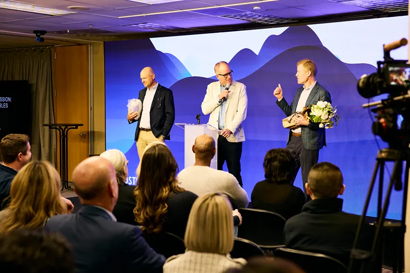 Three men on stage at a corporate event, one receiving an award with flowers, while another speaks at a podium to an audience.