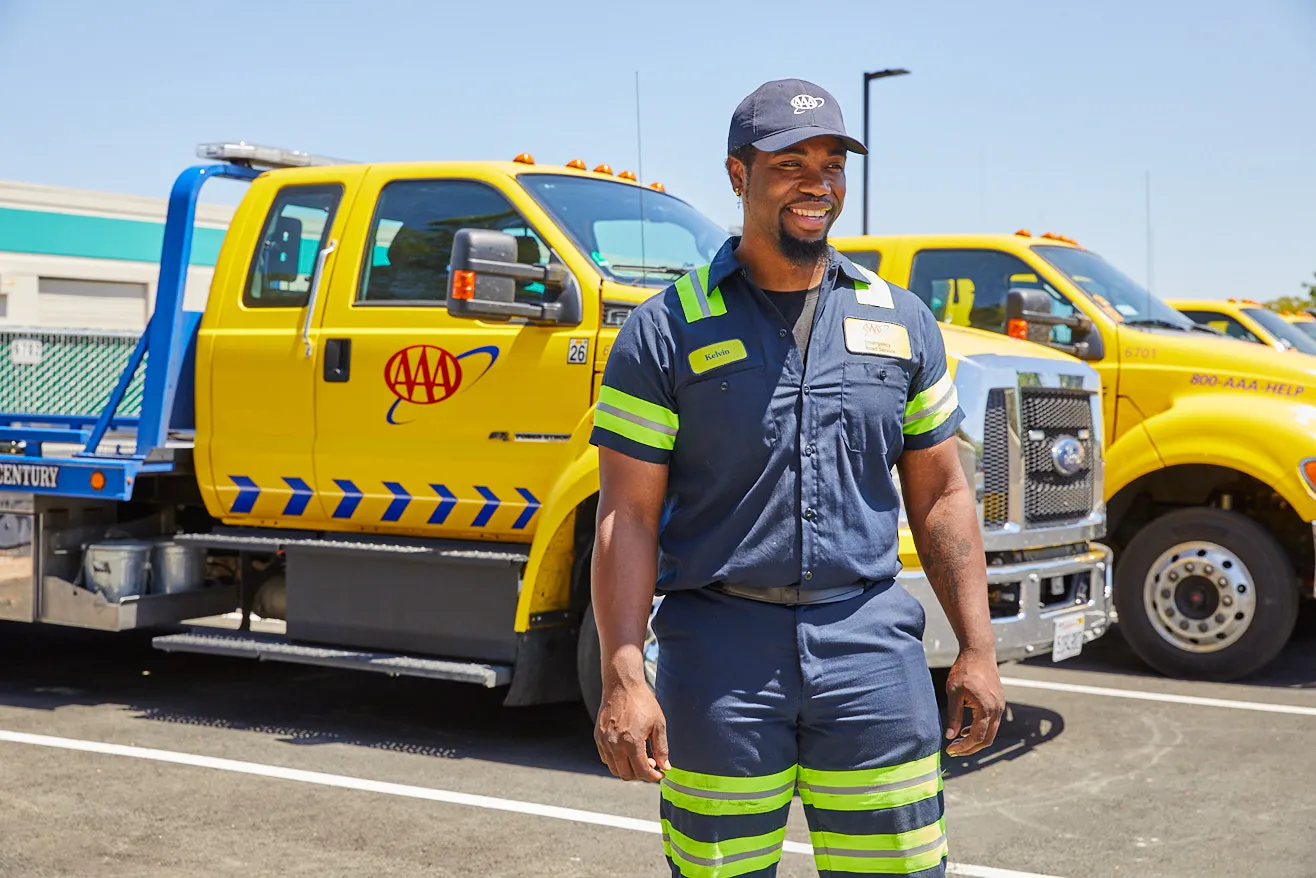 A smiling worker in uniform stands near yellow tow trucks under clear skies.