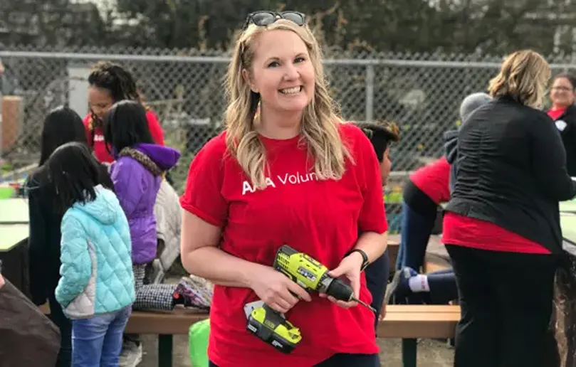 Woman in red shirt holding drill smiling at camera with others working behind her