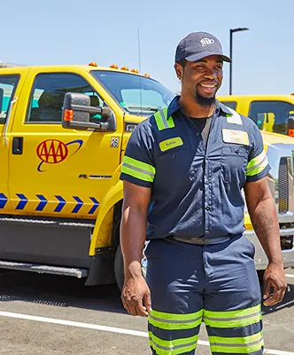 A smiling man in uniform stands near AAA tow trucks on a sunny day.