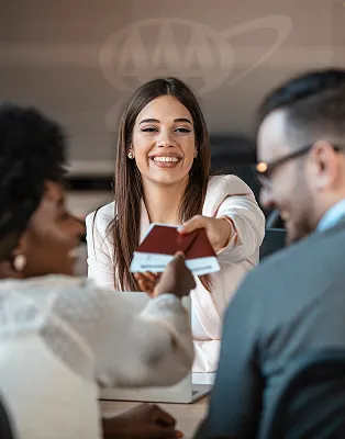A smiling woman hands passports to two people at a table.