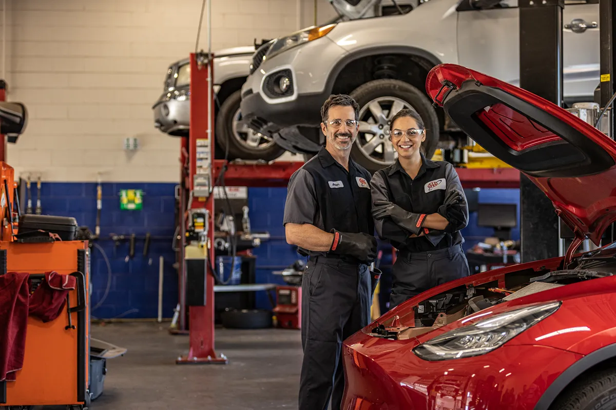 Two mechanics stand by red car with hood open in garage setting