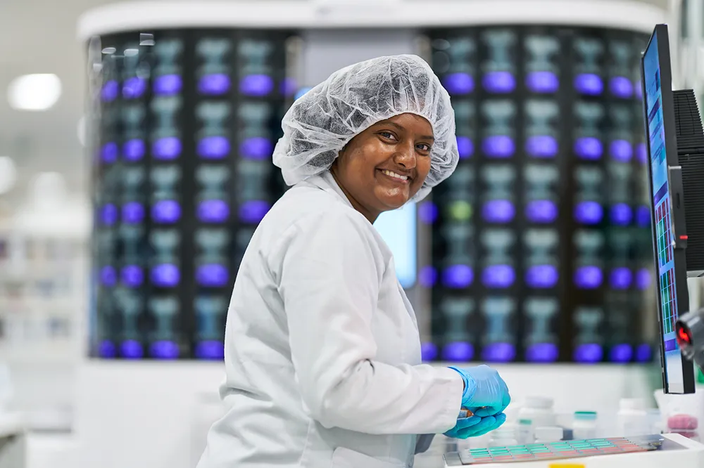A smiling healthcare professional is looking over her shoulder at the camera in a modern lab setting.