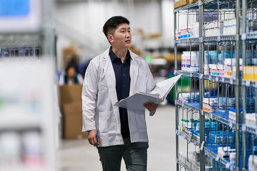 A male pharmacist in a lab coat is inspecting inventory on a shelf in a pharmacy.