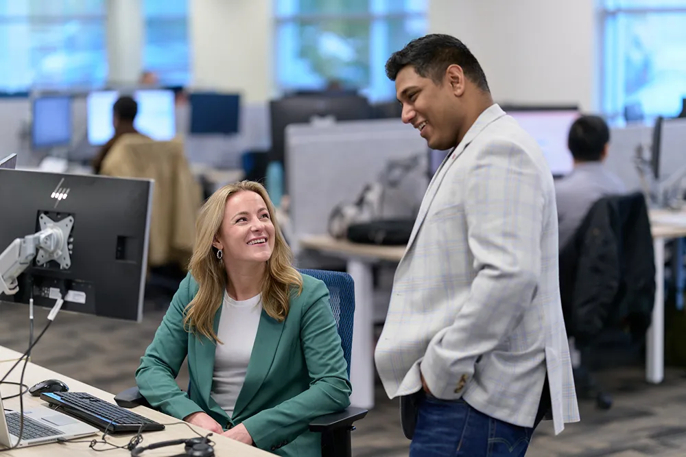 Two friendly corporate professionals are smiling while discussing a project at a desk in an office